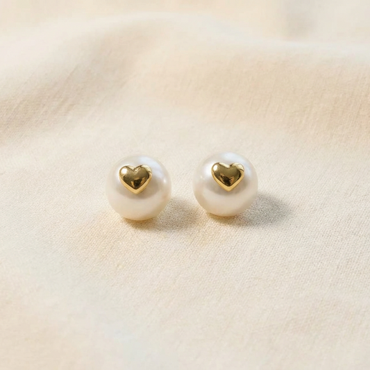 The Golden Heart pearl stud earrings with freshwater pearls and polished gold-plated heart detail on a warm beige fabric background