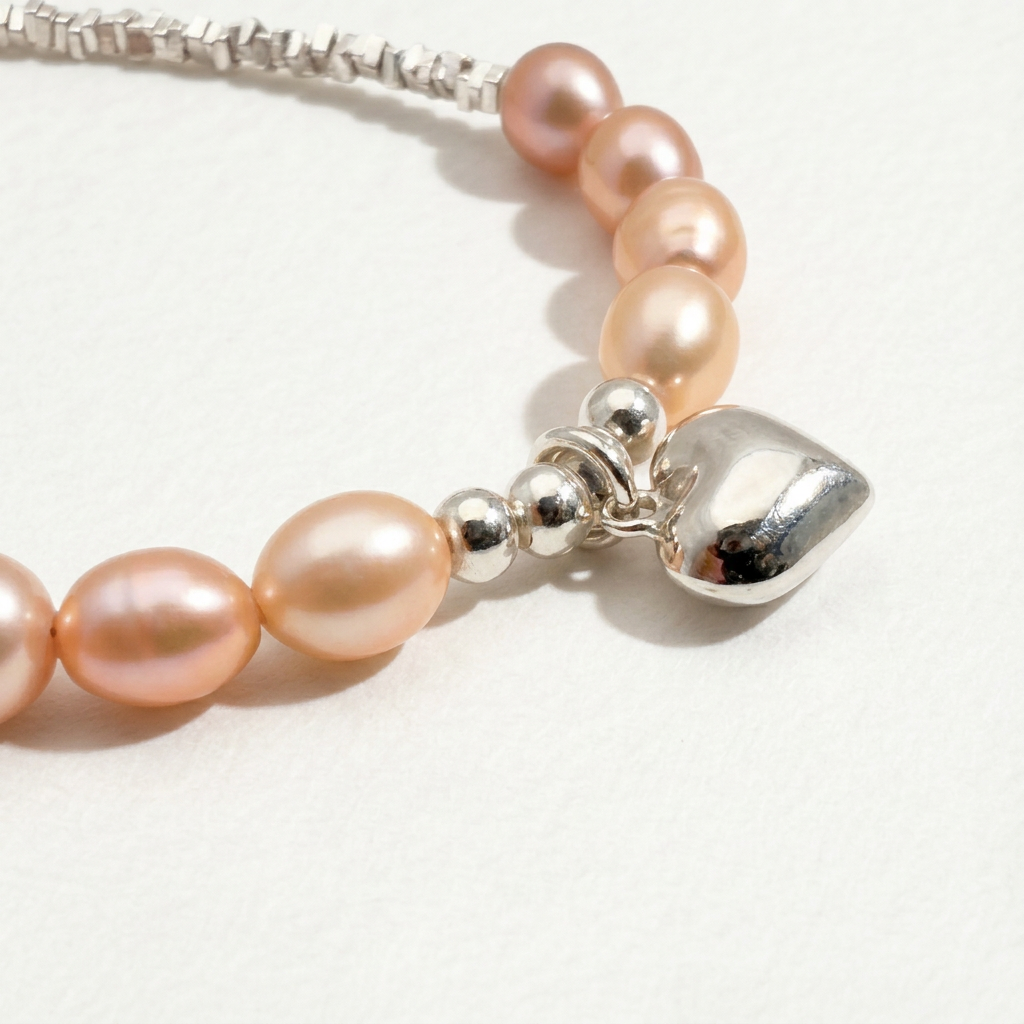 Close-up of The Soft Heart bracelet showing peach freshwater pearls, sterling silver beads, and a heart charm detail on a light beige background