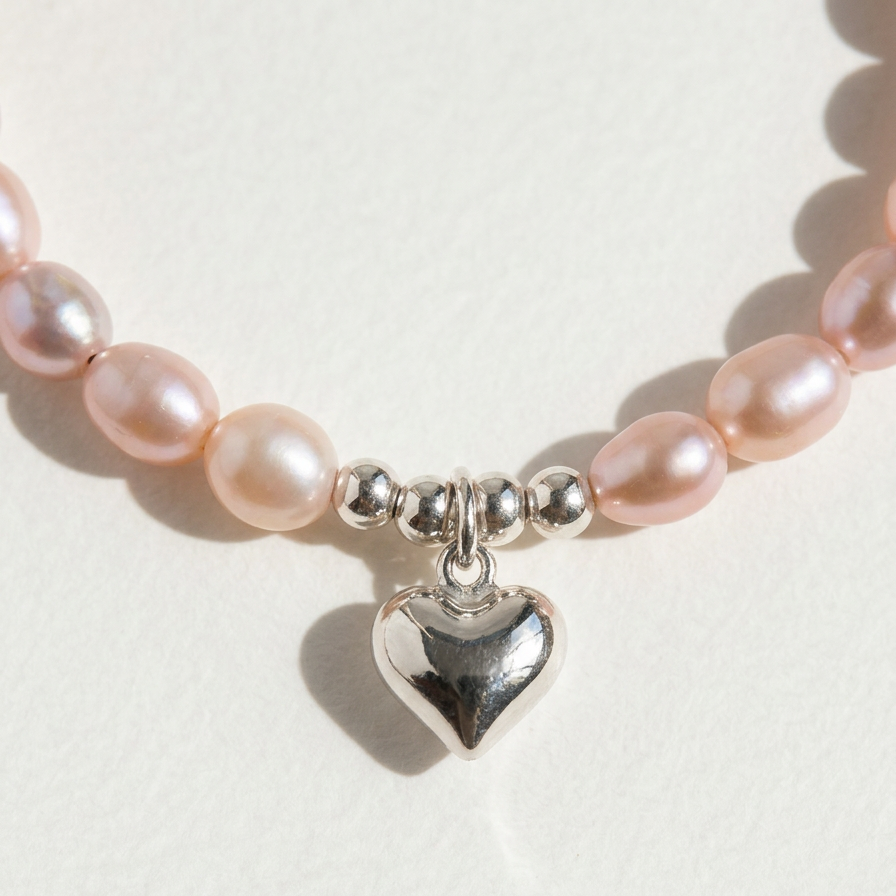 Close-up of The Soft Heart bracelet showing peach freshwater pearls, sterling silver beads, and a heart charm on a light beige background